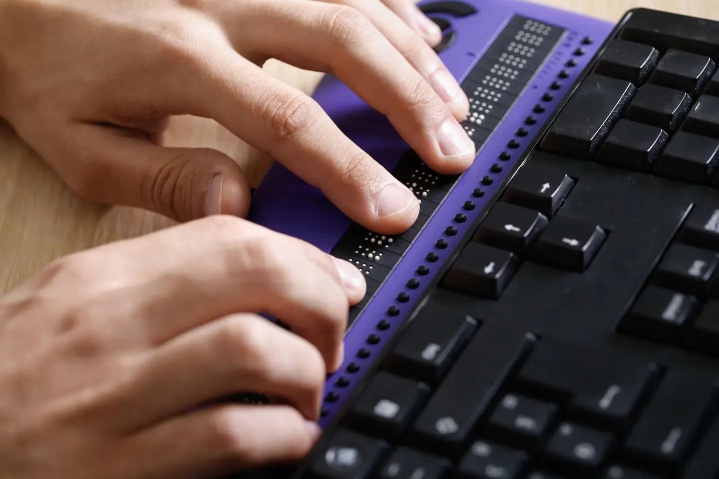 Hands using a Braille display next to a keyboard on a wooden table.