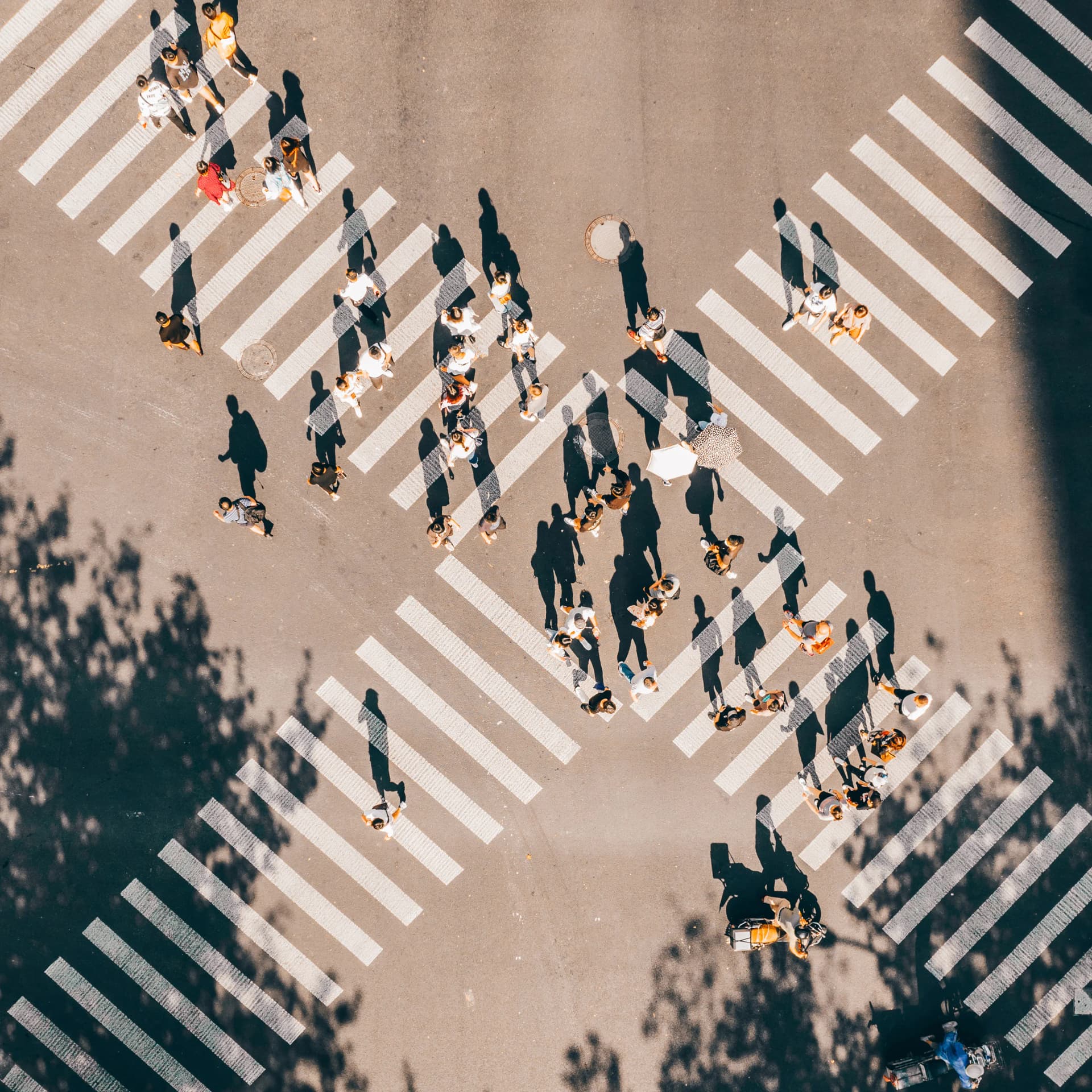 Aerial view of people crossing intersecting crosswalks on a sunny day, casting long shadows on the street.