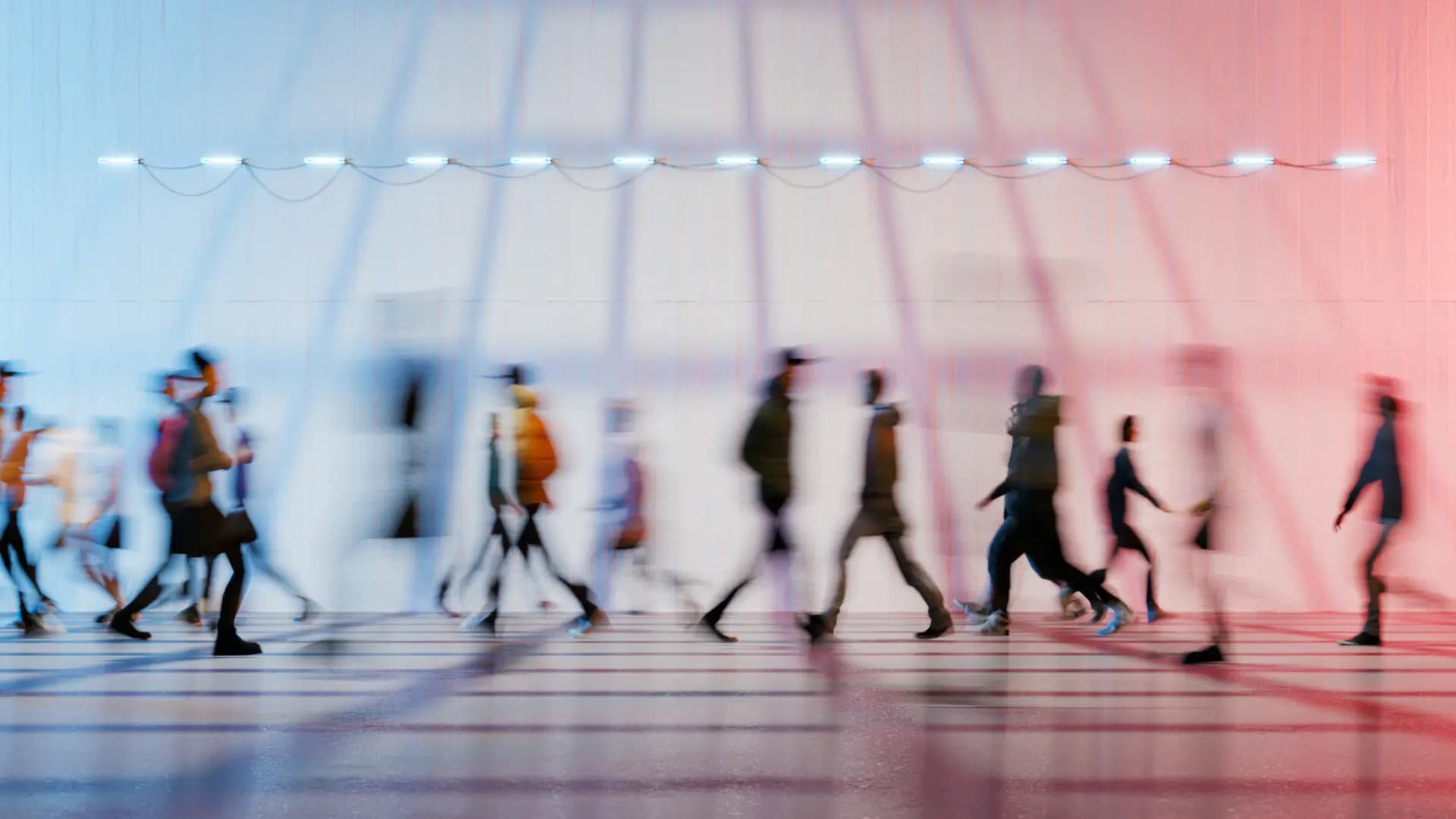 Blurred image of people walking in front of a white wall with red and blue lighting, casting long shadows.