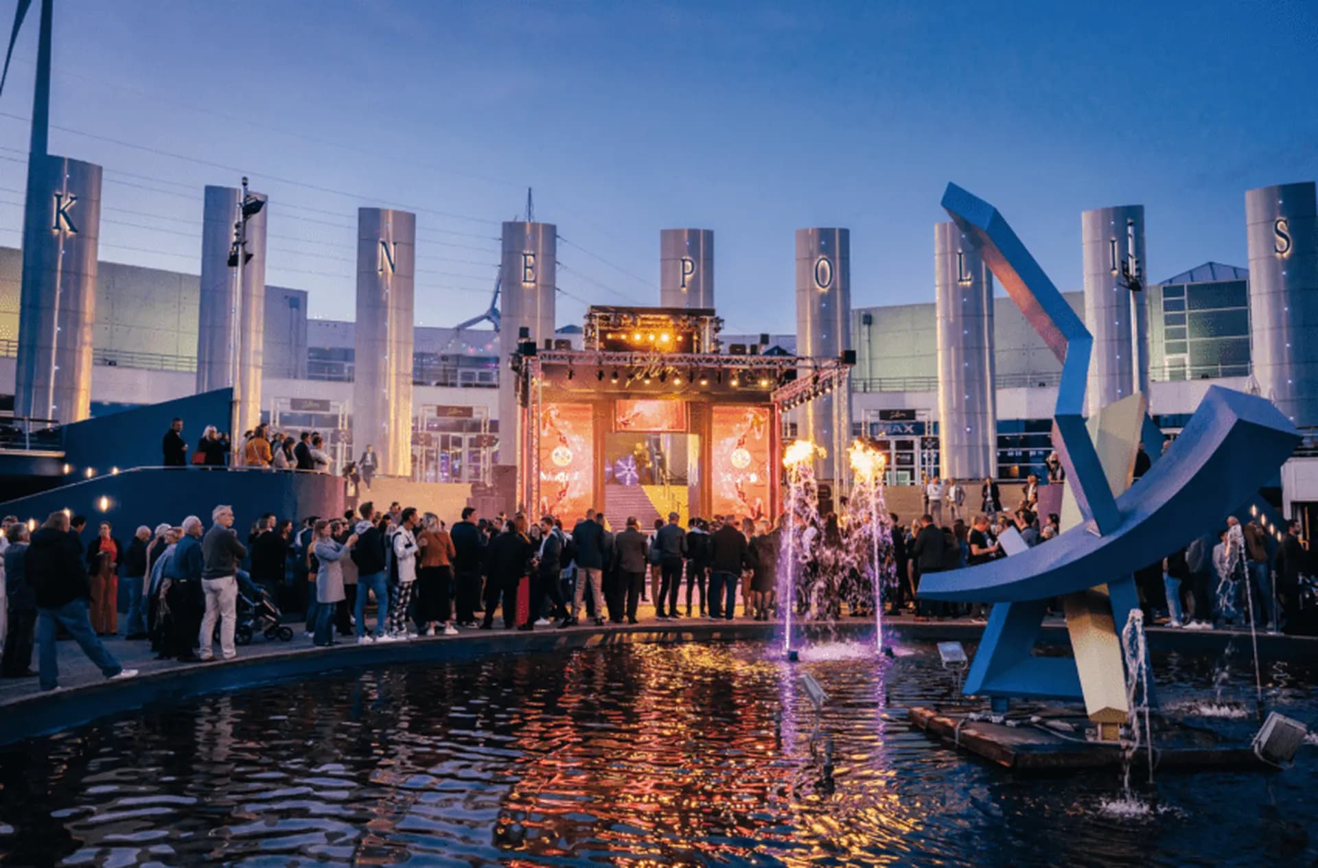 Crowd gathers at an outdoor event with colorful lights, a stage, and modern sculptures reflecting in a nearby pond at dusk.