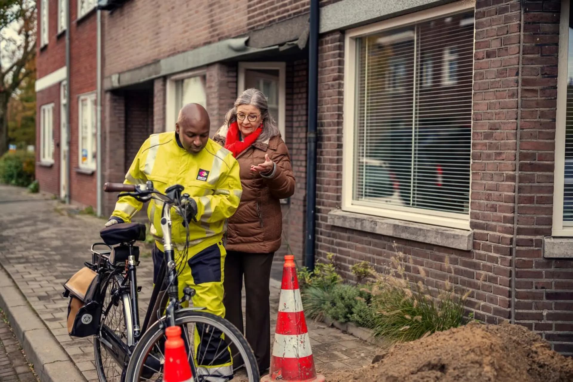 A person in a bright yellow uniform with a bicycle stands next to a woman in a brown jacket. They appear to be conversing near traffic cones.