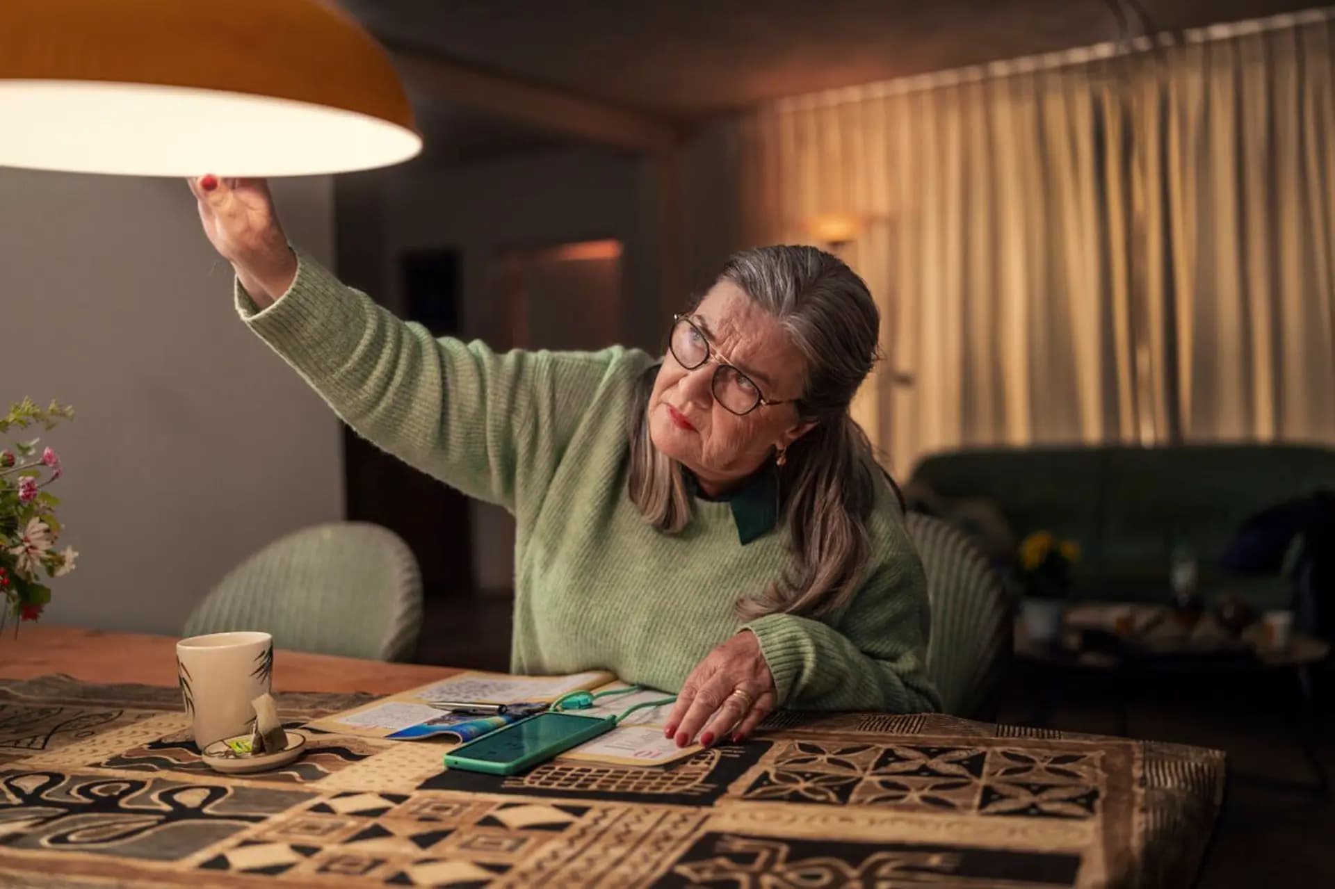 Older woman in glasses adjusts a hanging lamp above a table with a patterned cloth, a cup, and a smartphone. She looks focused.