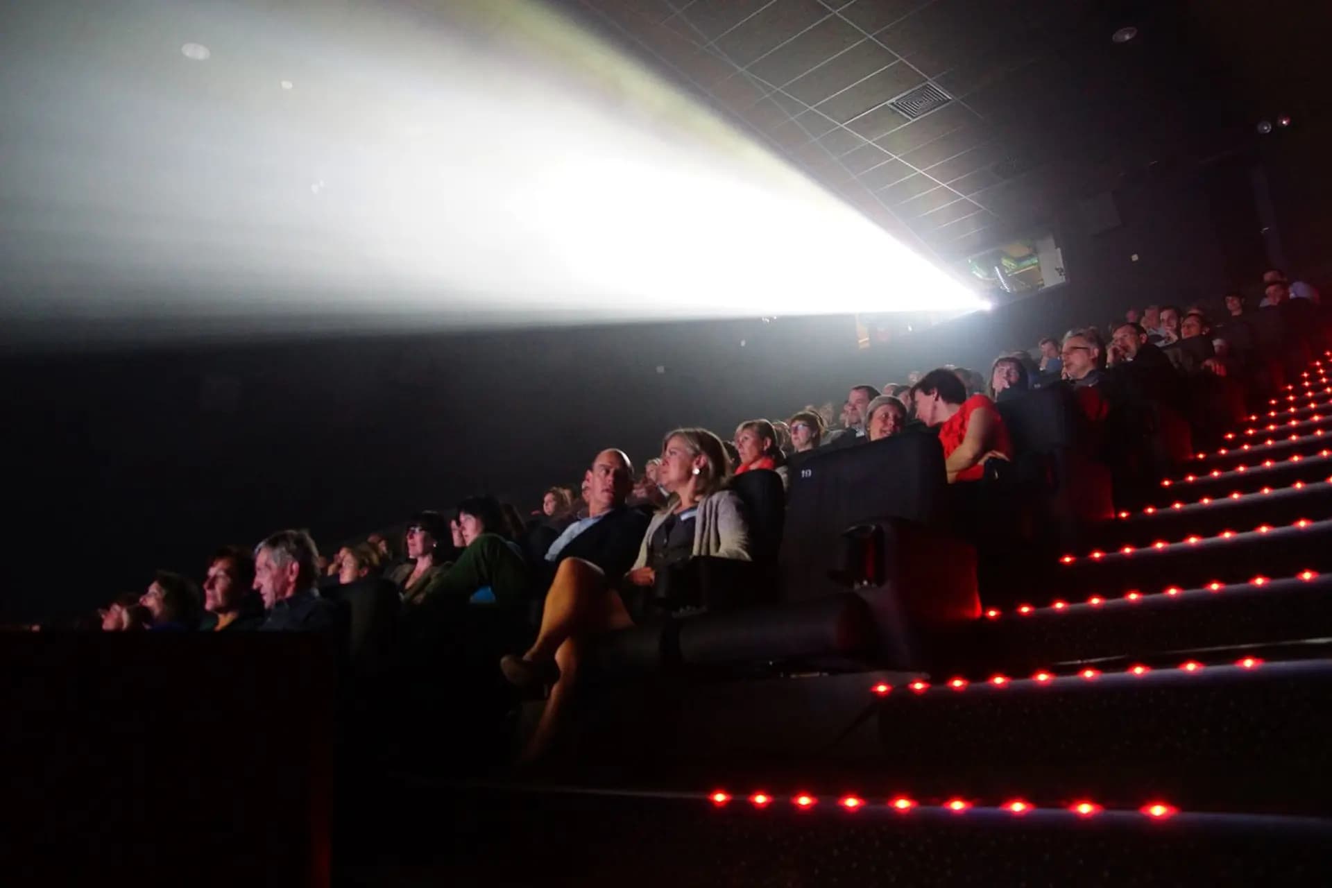 Audience seated in a dark theater with a bright projector beam illuminating the scene, and red aisle lights highlighting the stairs.