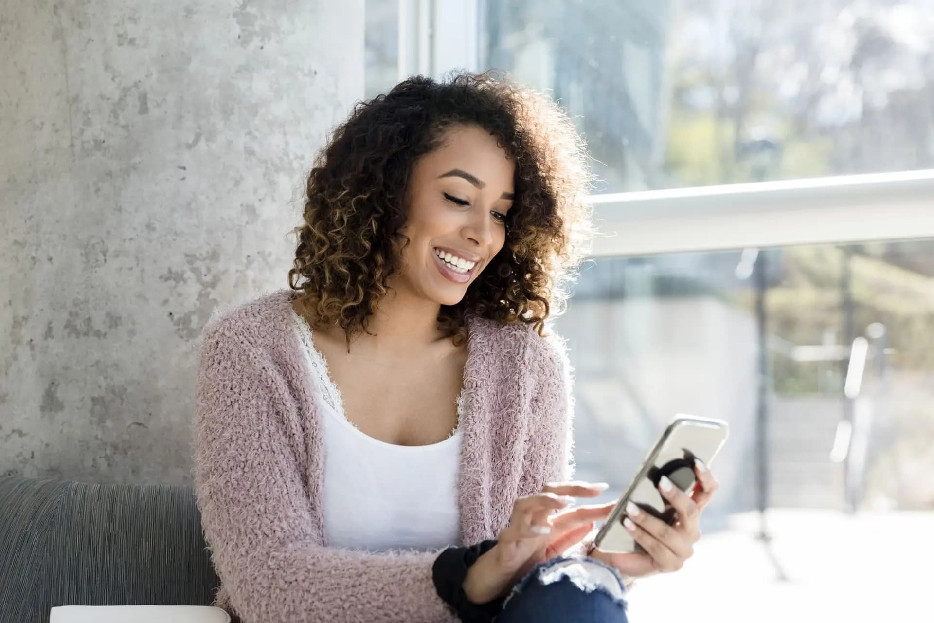 A woman with curly hair smiles while looking at her phone, sitting indoors with natural light streaming through a window.