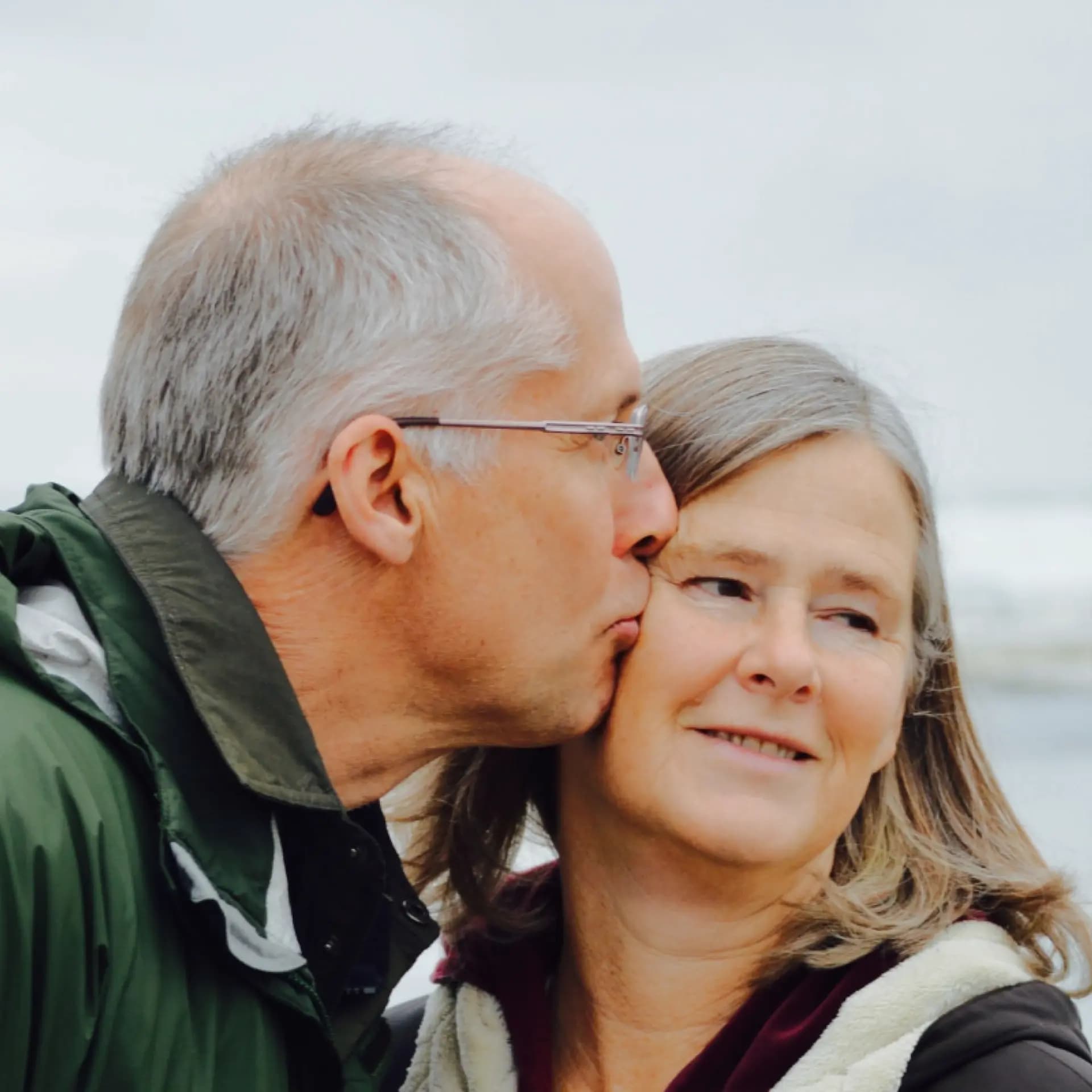 A man kisses a woman on the cheek outdoors. Both are wearing jackets. The woman smiles softly, and the background is blurred.