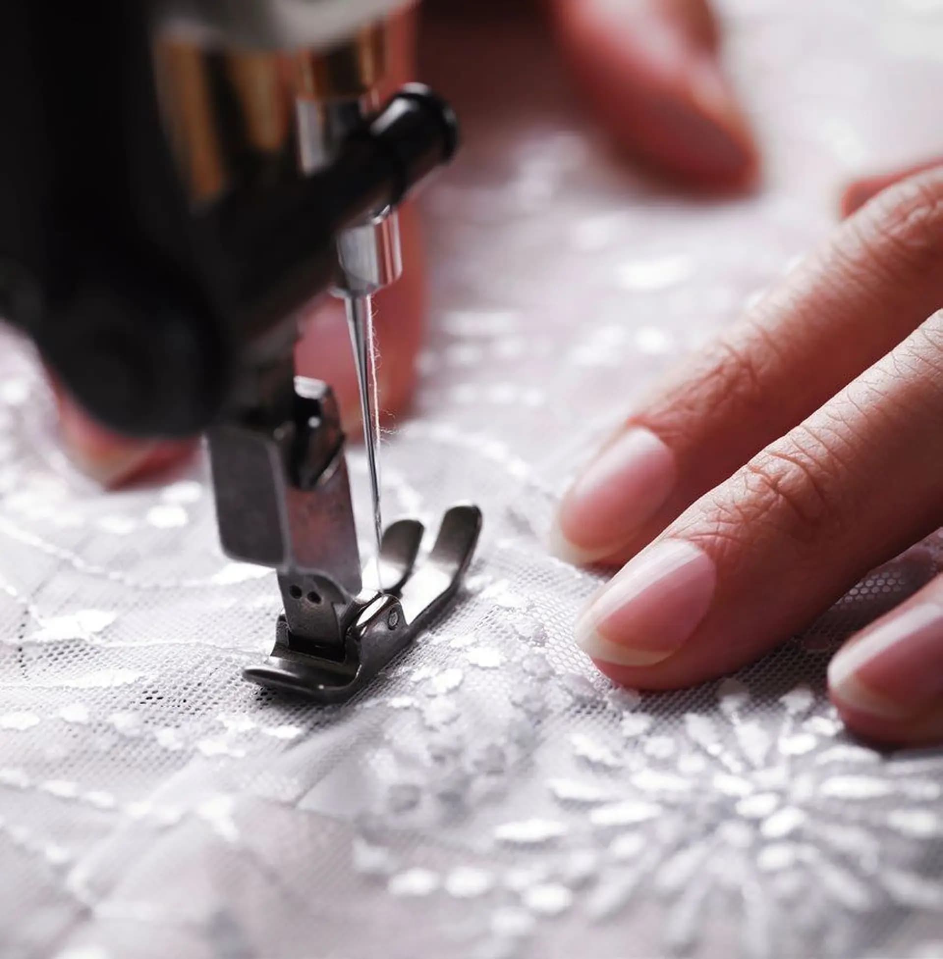 Close-up of a sewing machine needle stitching delicate white lace fabric, with a person's hand guiding the material.