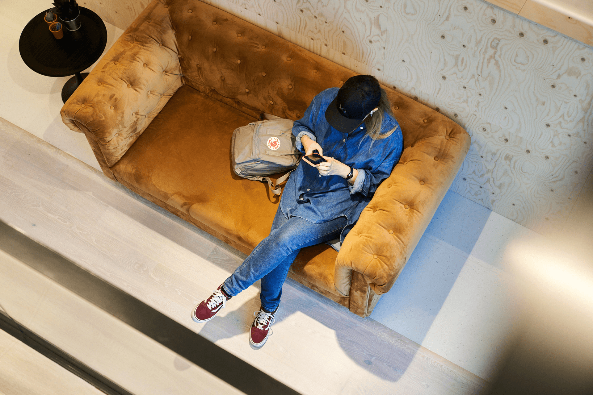 Person in denim outfit and black cap sits on a brown sofa, using a phone, with a backpack beside them. Top-view perspective.
