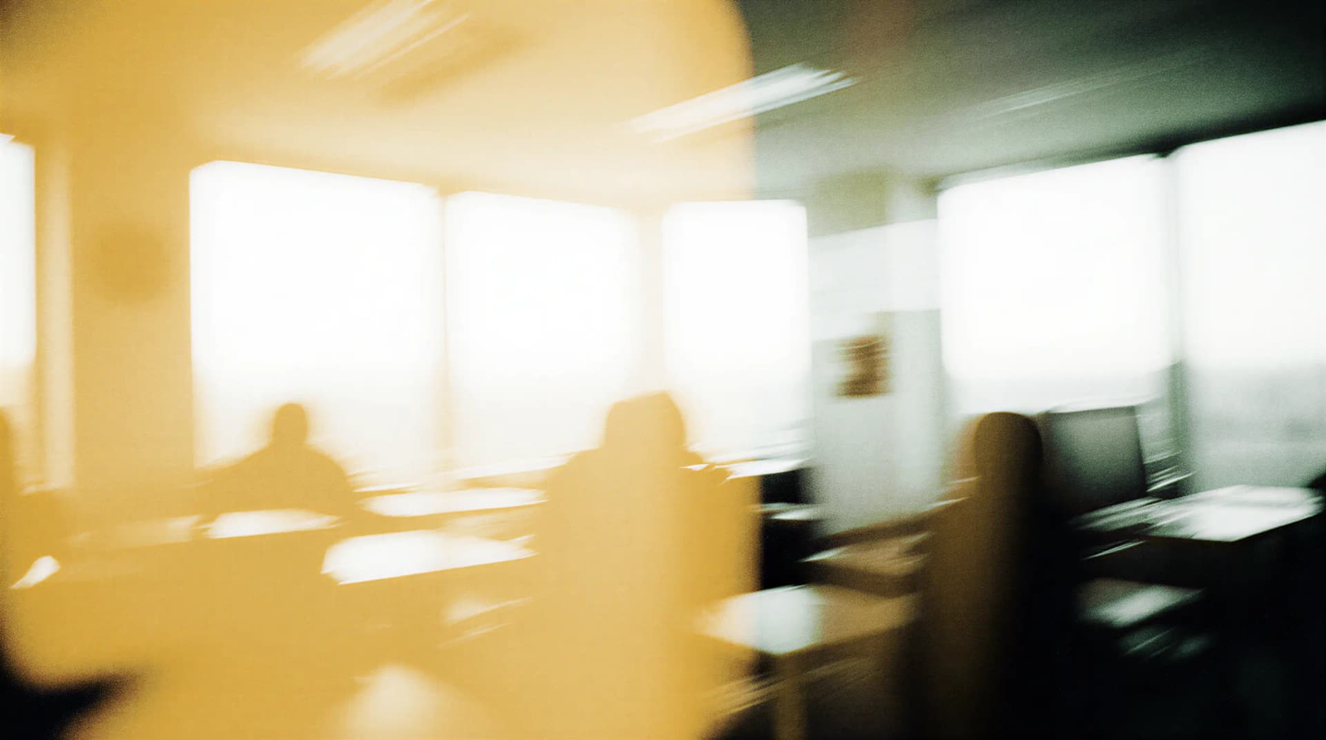 Blurry image of a sunlit room with silhouettes of people and furniture, creating an abstract and distorted view.