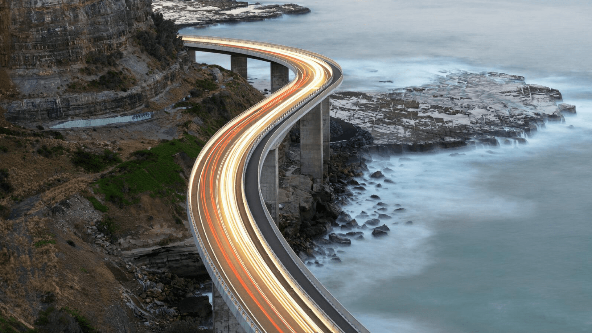 A coastal highway curves dramatically above rocky shores, with light trails from vehicles creating bright lines against the evening backdrop.