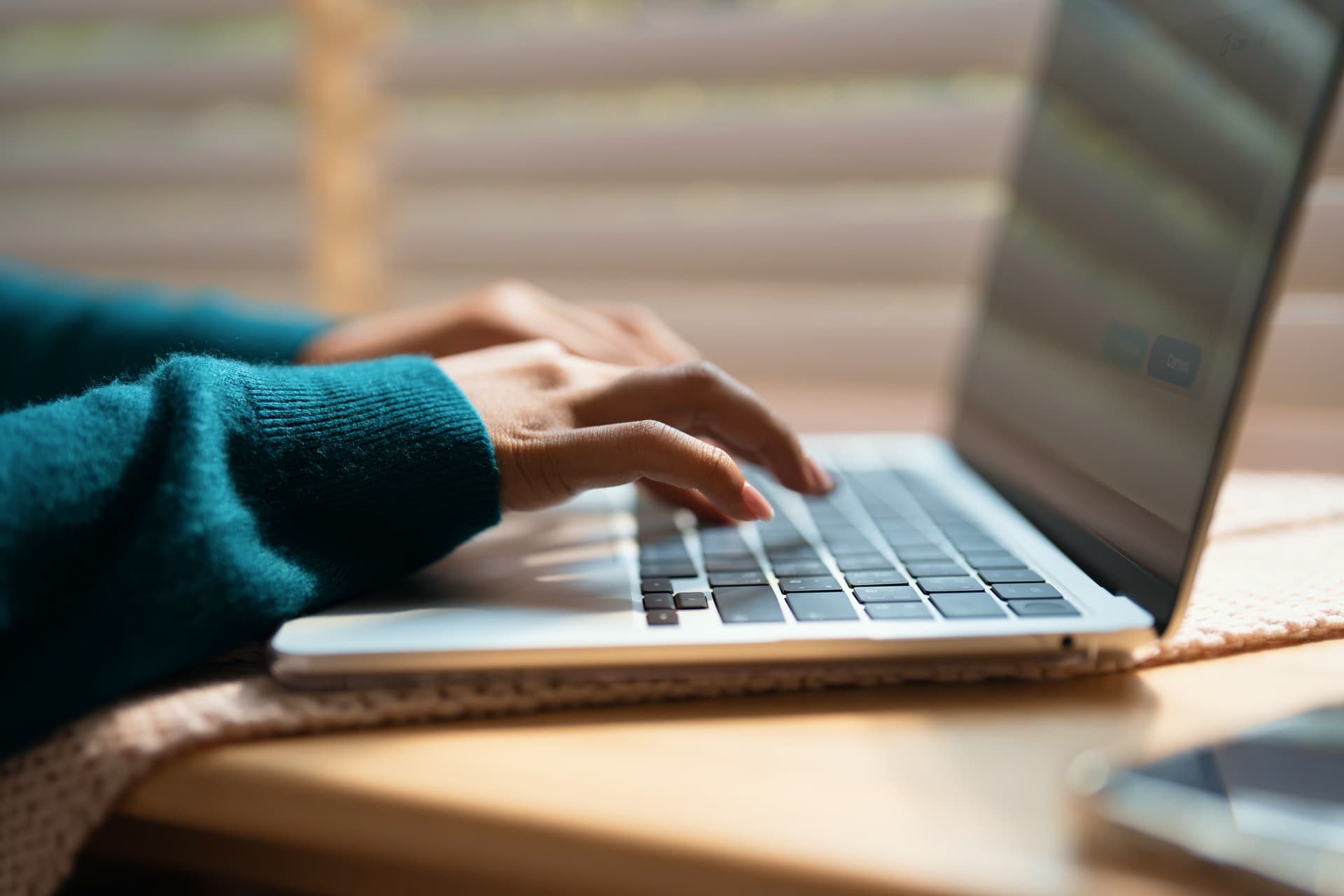 Hands typing on a laptop keyboard, with a focus on fingers pressing keys. The person is wearing a teal sweater, and blinds are visible in the background.