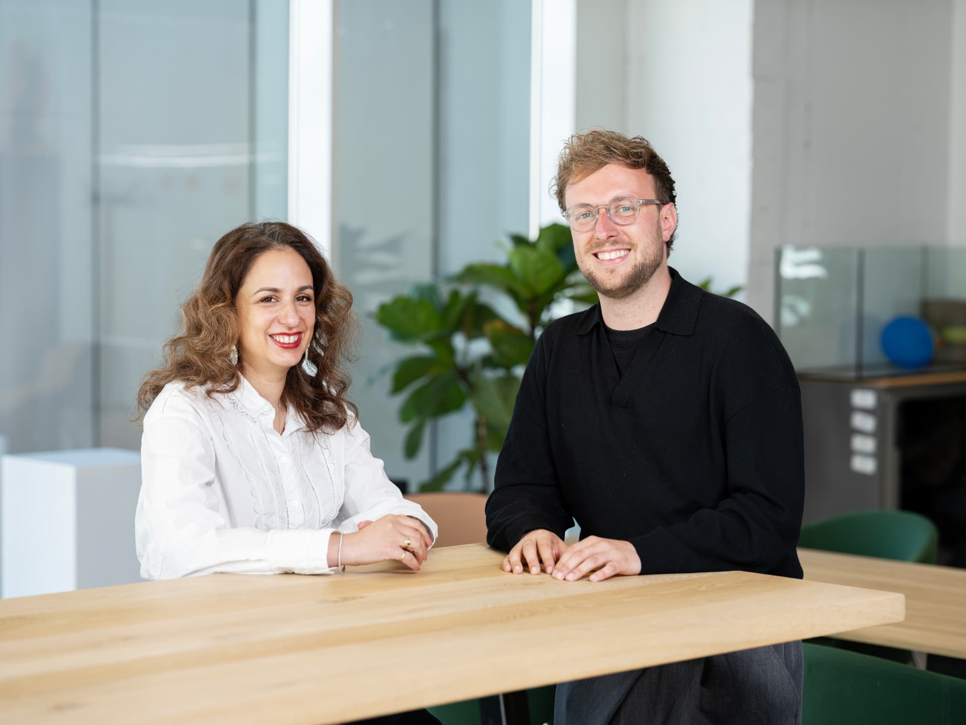 Two people smiling while sitting at a wooden table in a modern office setting with a plant in the background.