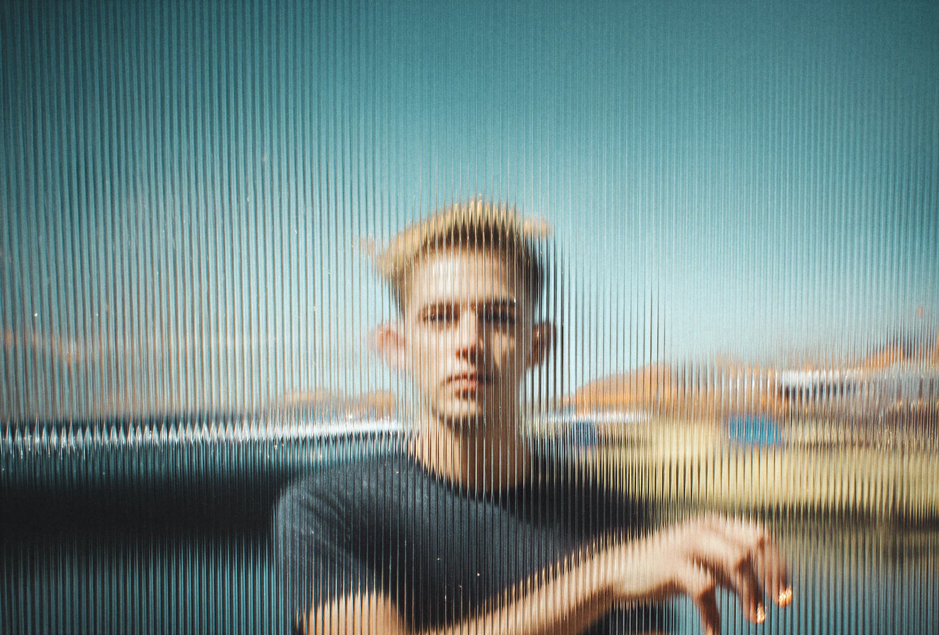 Blurred person behind a textured glass, with vertical lines creating a distorted effect against a blue sky background.