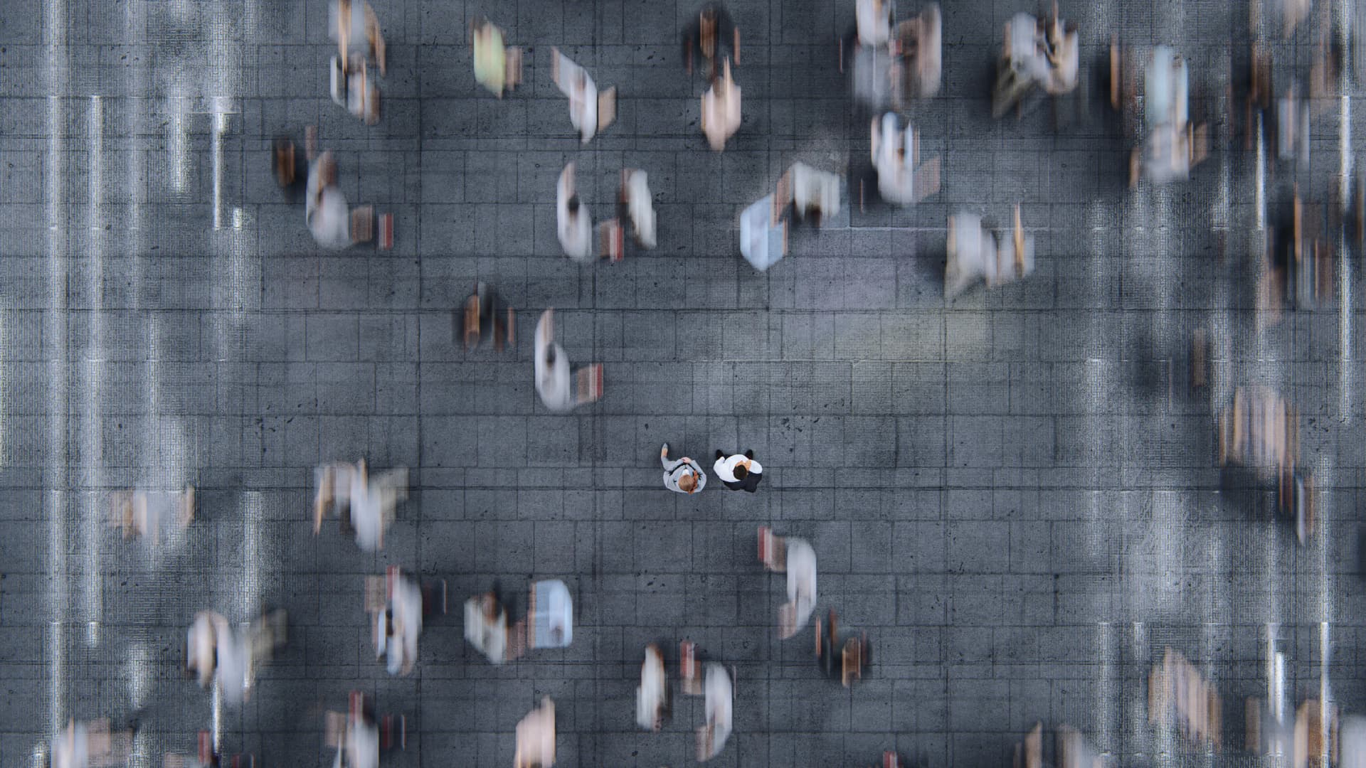 Aerial view of a busy, blurred crowd on a pavement, with two clear individuals standing in the center, creating a contrast.