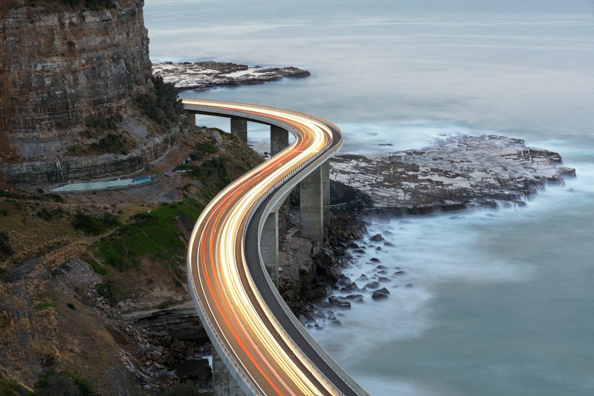 Curved coastal bridge with light trails, adjacent to cliffs and ocean waves below, under a cloudy sky.