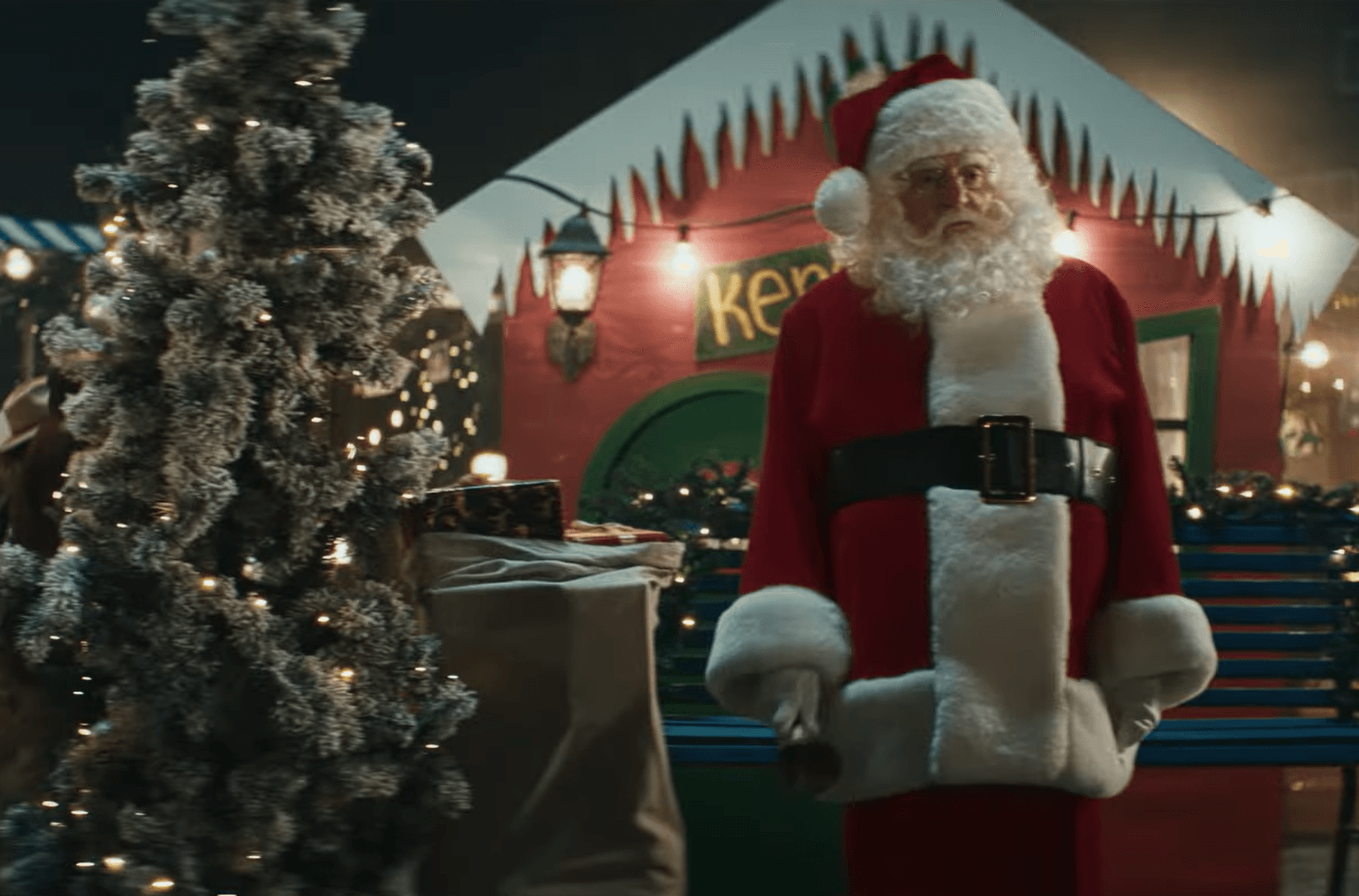 Santa in red suit and white beard stands beside a snow‑flocked Christmas tree, wrapped gifts and a festive red holiday hut.