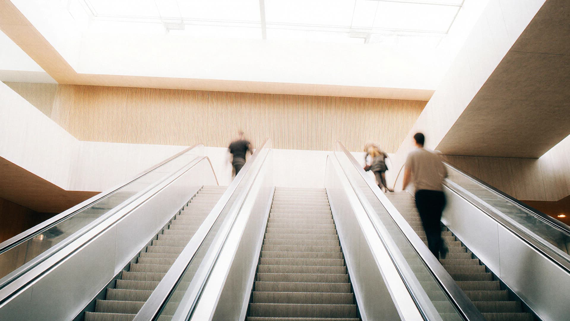 Three people ascending two parallel escalators in a modern, brightly lit building interior.