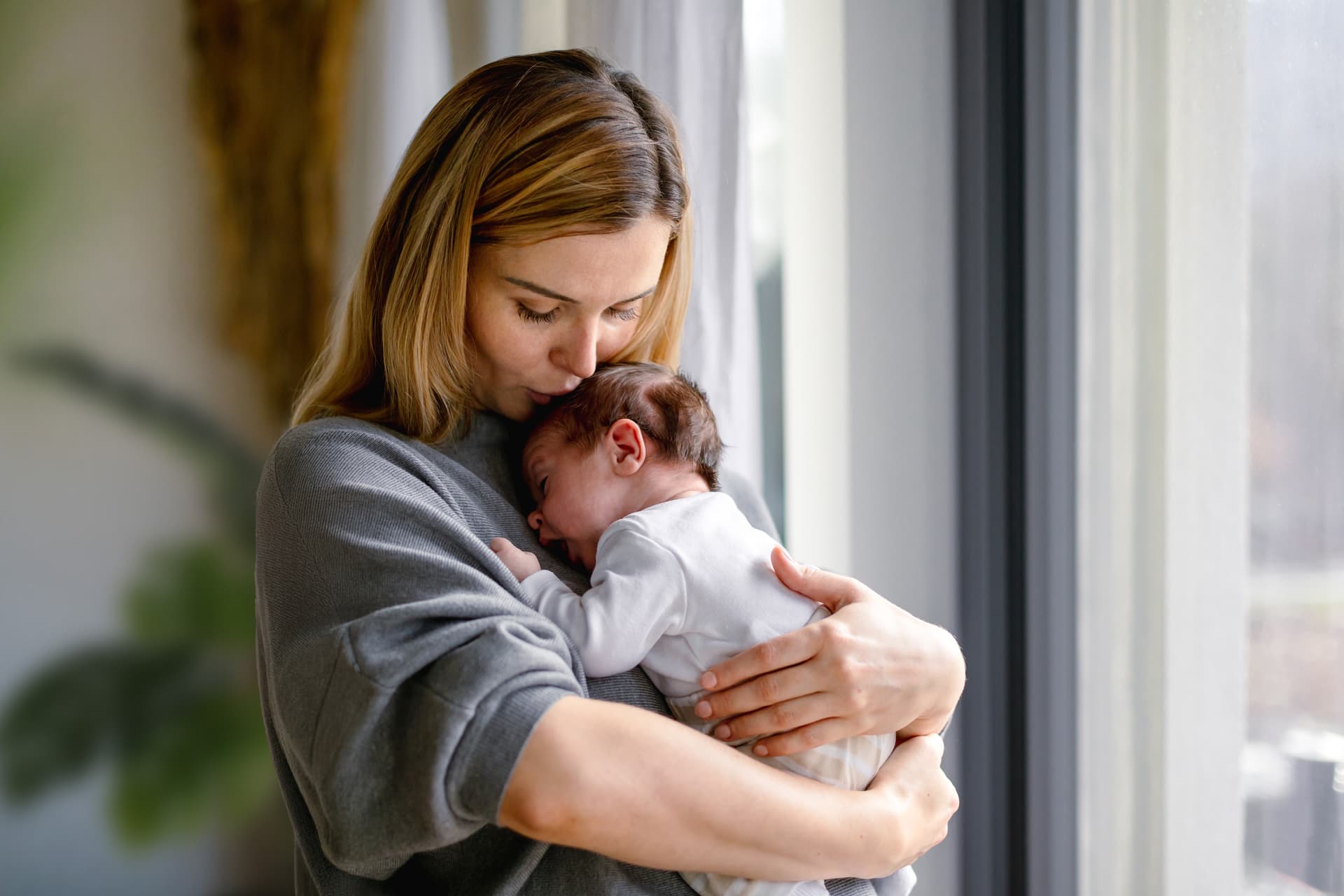 A woman gently holding and kissing a newborn baby, standing by a window with soft natural light filtering in.