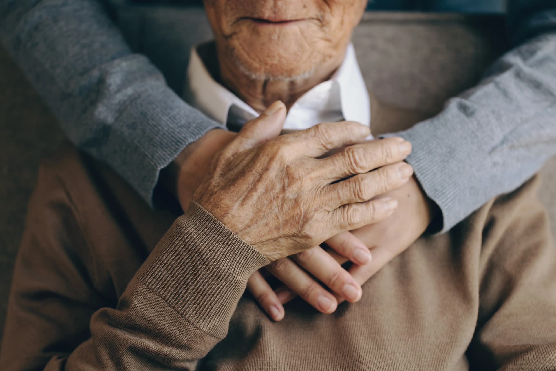 An elderly person in a brown sweater is embraced warmly by another person with hands resting gently on their chest.