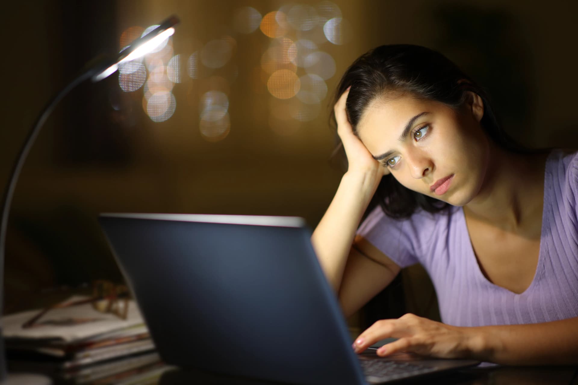 Woman in a purple shirt, looking tired and leaning on her hand, works on a laptop under a desk lamp with blurred lights in the background.