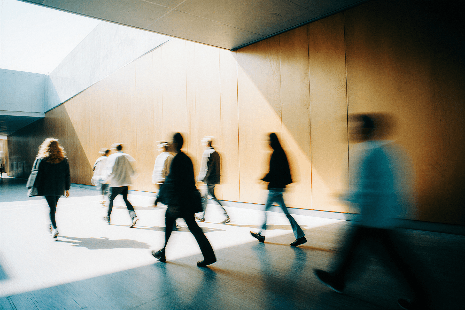 Blurred figures walking in a sunlit hallway with wooden walls, creating a sense of motion and bustling activity.