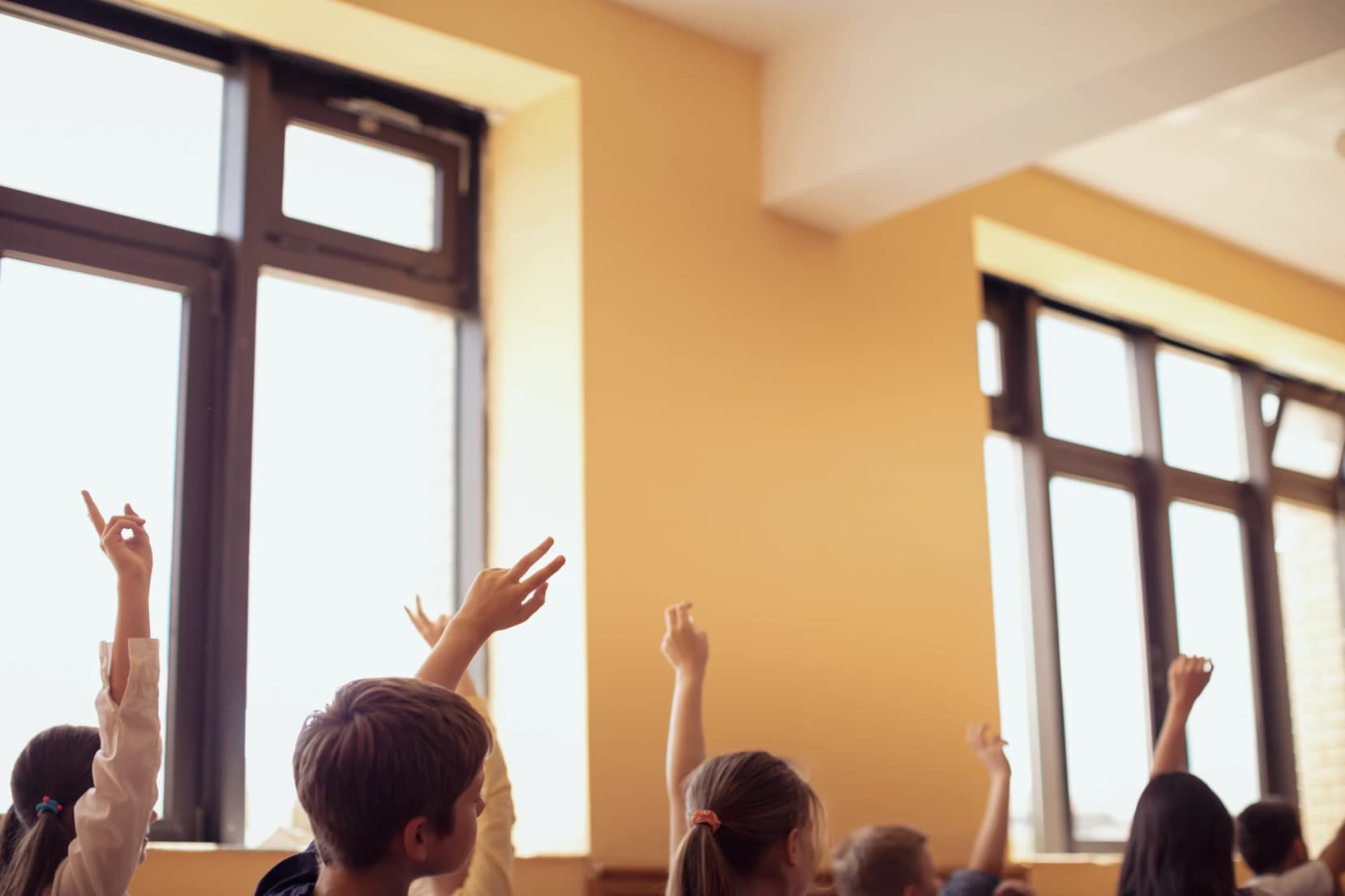 Students in a classroom with yellow walls, raising their hands, possibly to answer a question or participate in a discussion.