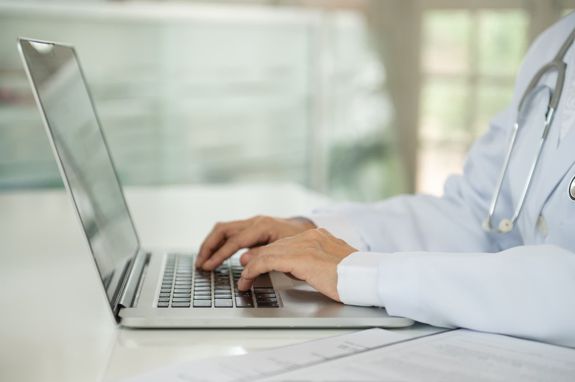 A doctor types on a laptop at a desk, with medical documents nearby, in a bright and modern setting.