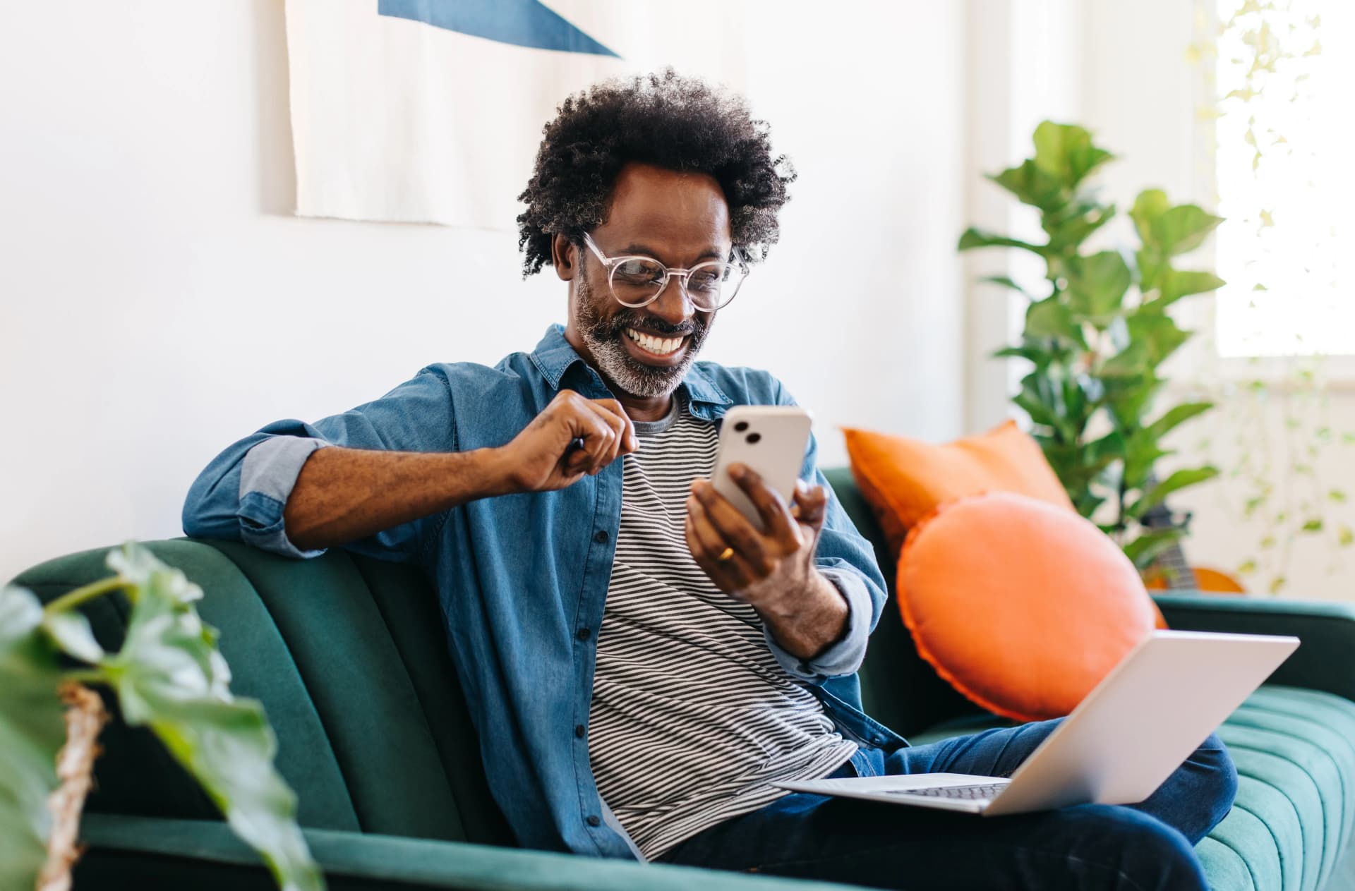 Smiling man with glasses using a smartphone, sitting on a green couch with a laptop and orange pillow, surrounded by plants.