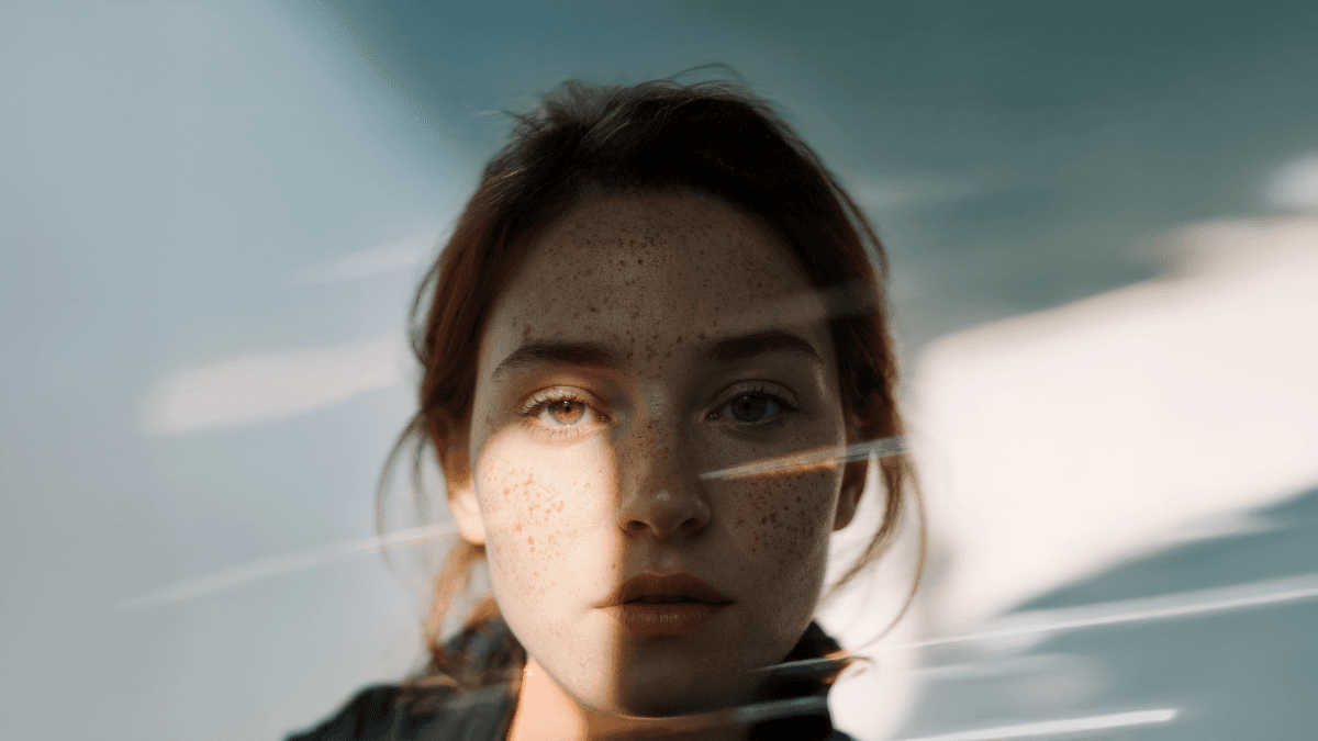 A woman with freckles is illuminated by sunlight, creating soft shadows on her face, with a blurred background.