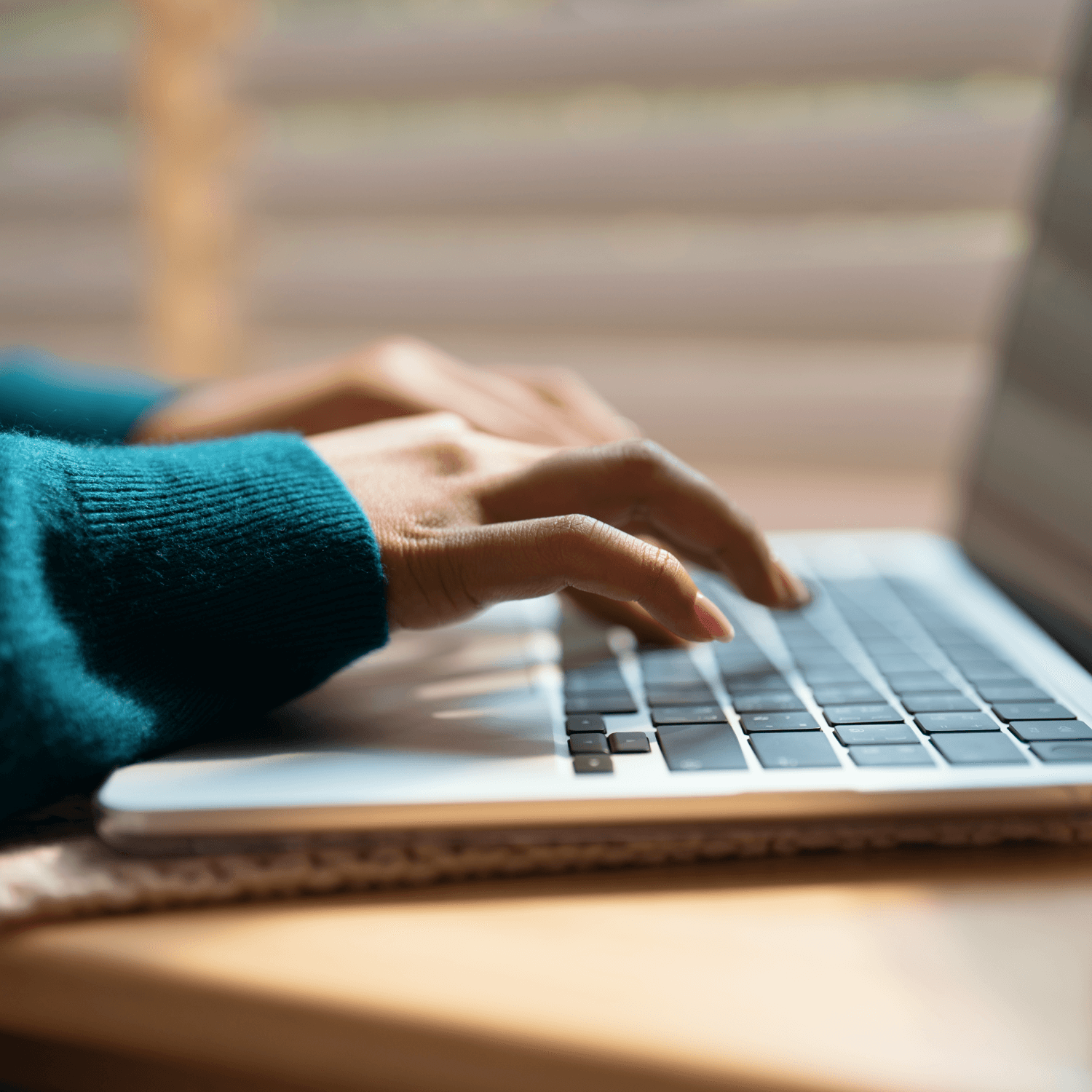 Close-up of hands typing on a laptop keyboard, with sunlight filtering through blinds in the background.