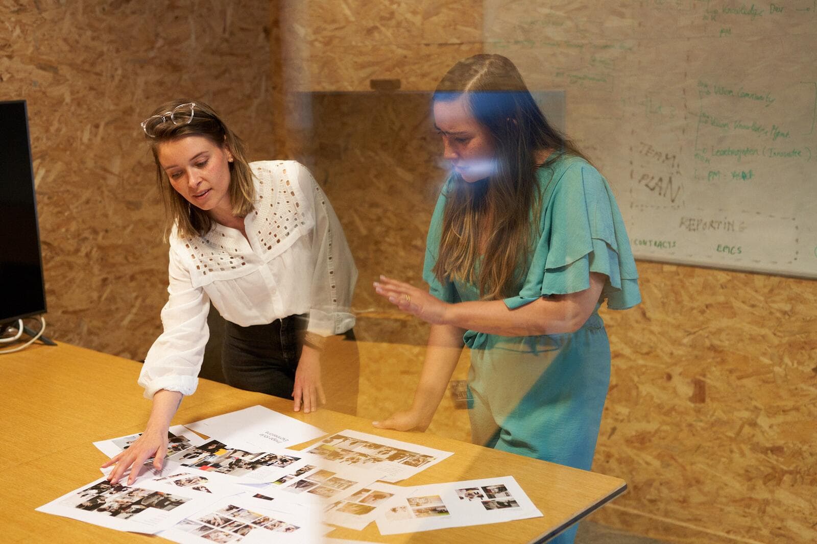 Two women stand at a table examining printed materials in a conference room. A whiteboard with notes is visible in the background.