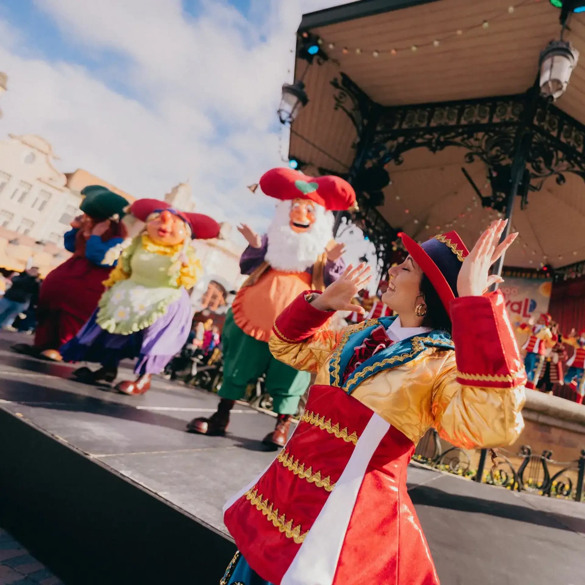Performers in colorful costumes dance energetically on a stage during an outdoor festival, with a festive pavilion in the background.