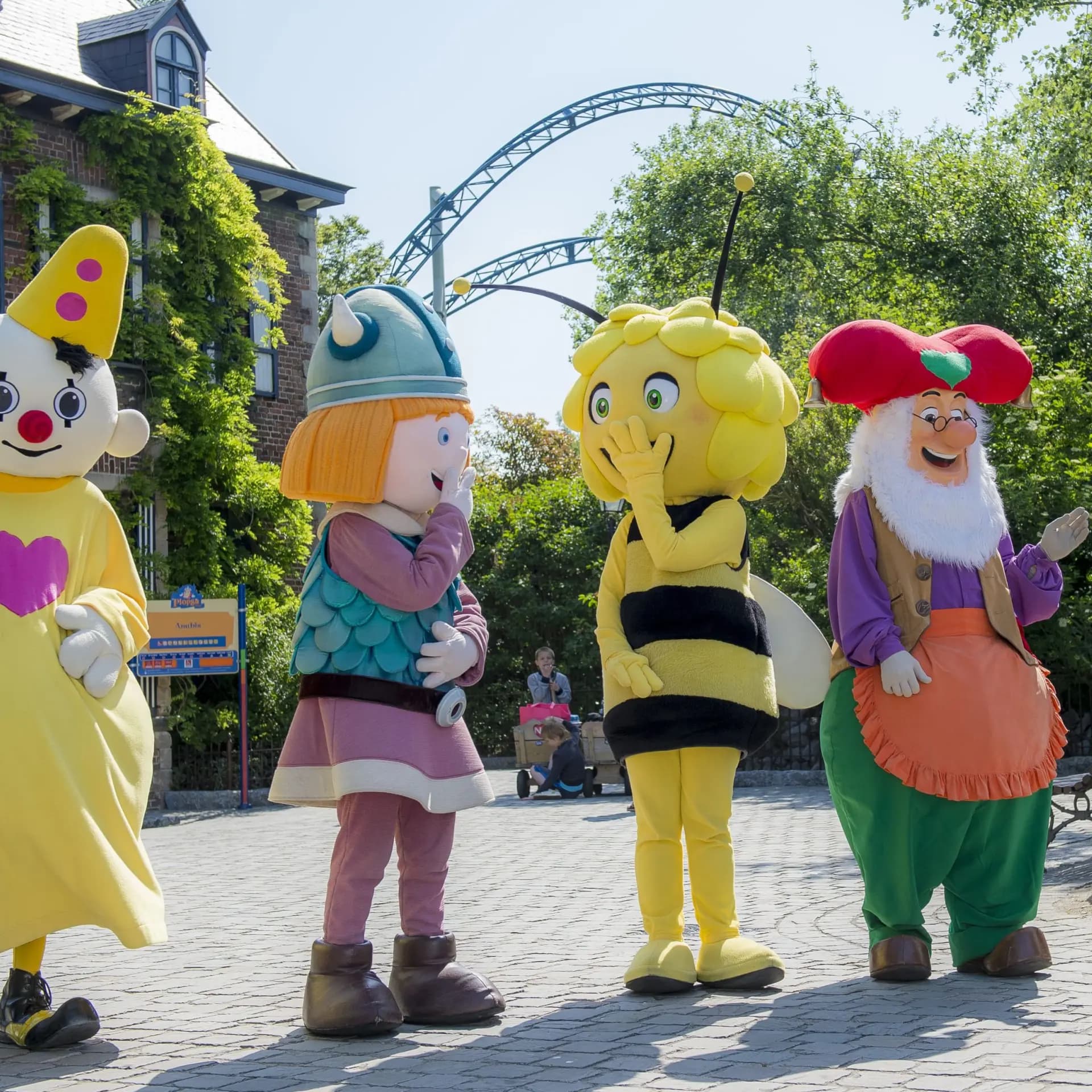 Four colorful mascots, including a clown, a viking, a bee, and a dwarf, standing on a cobblestone path with a roller coaster in the background.