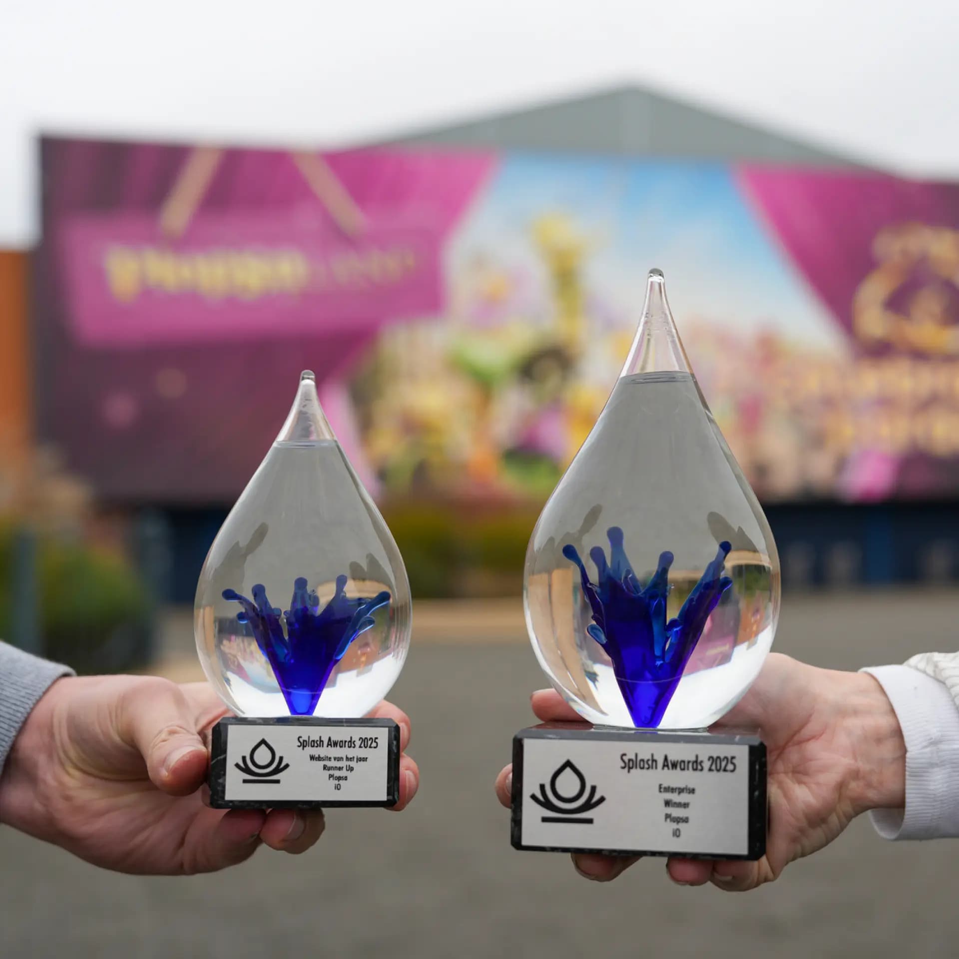 Two hands hold glass drop-shaped Splash Awards 2023 trophies, with blue splashes inside, against a blurred colorful background.
