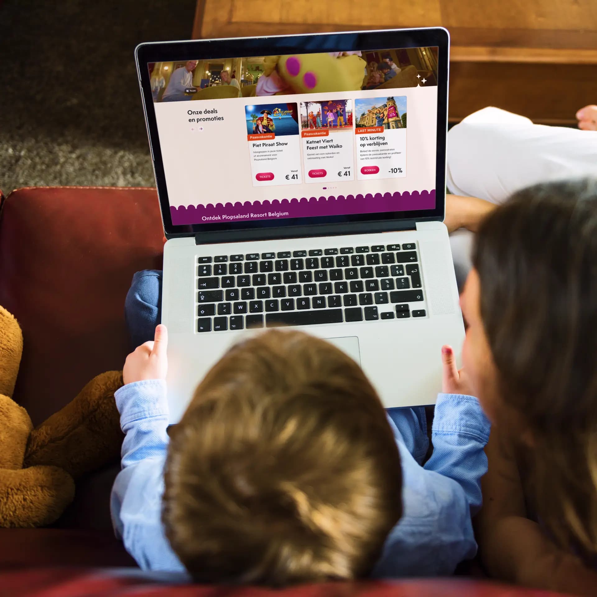 A child and an adult looking at amusement park deals on a laptop while sitting on a couch, with a teddy bear nearby.