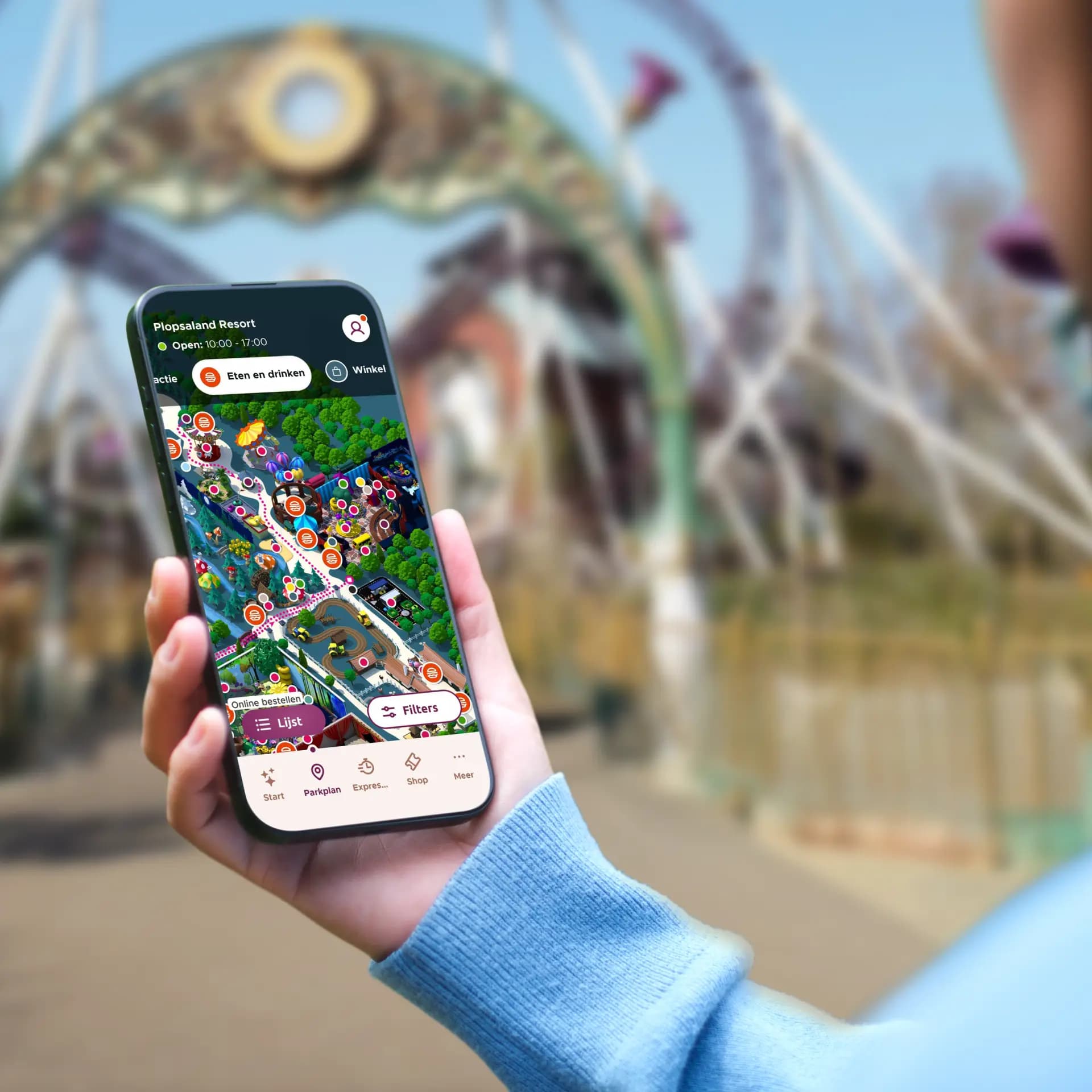 Person holding a smartphone displaying a colorful amusement park map app, with a blurred park entrance archway in the background.