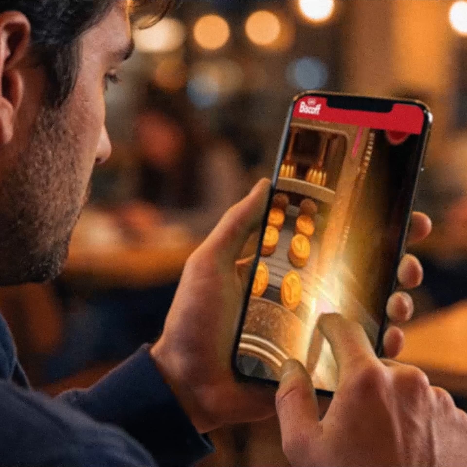A person holding a smartphone, playing a coin-slot game with a finger tapping the screen, in a warmly lit indoor setting.