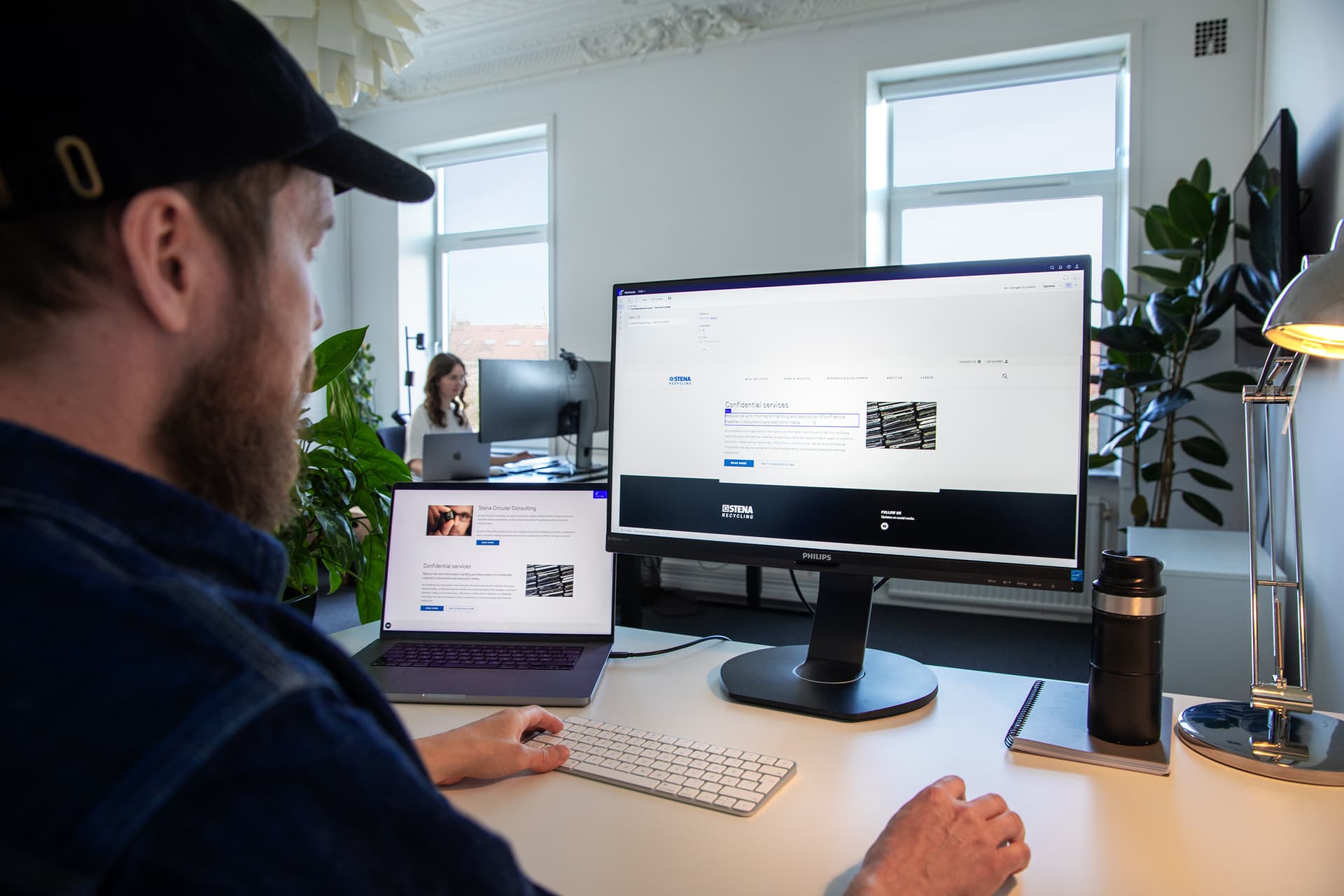 A person in a cap working at a desk with a computer monitor and laptop. Another person works in the background in a bright office with plants.