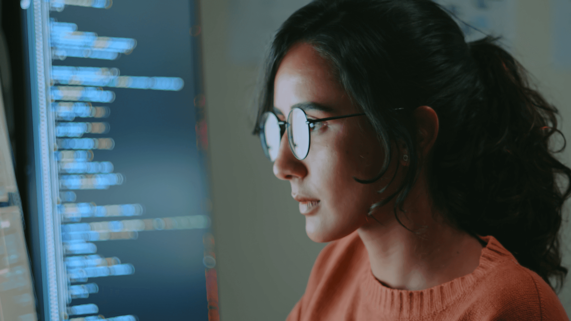 A woman with glasses focused on coding on a computer screen, highlighting lines of code, in a dimly lit setting.