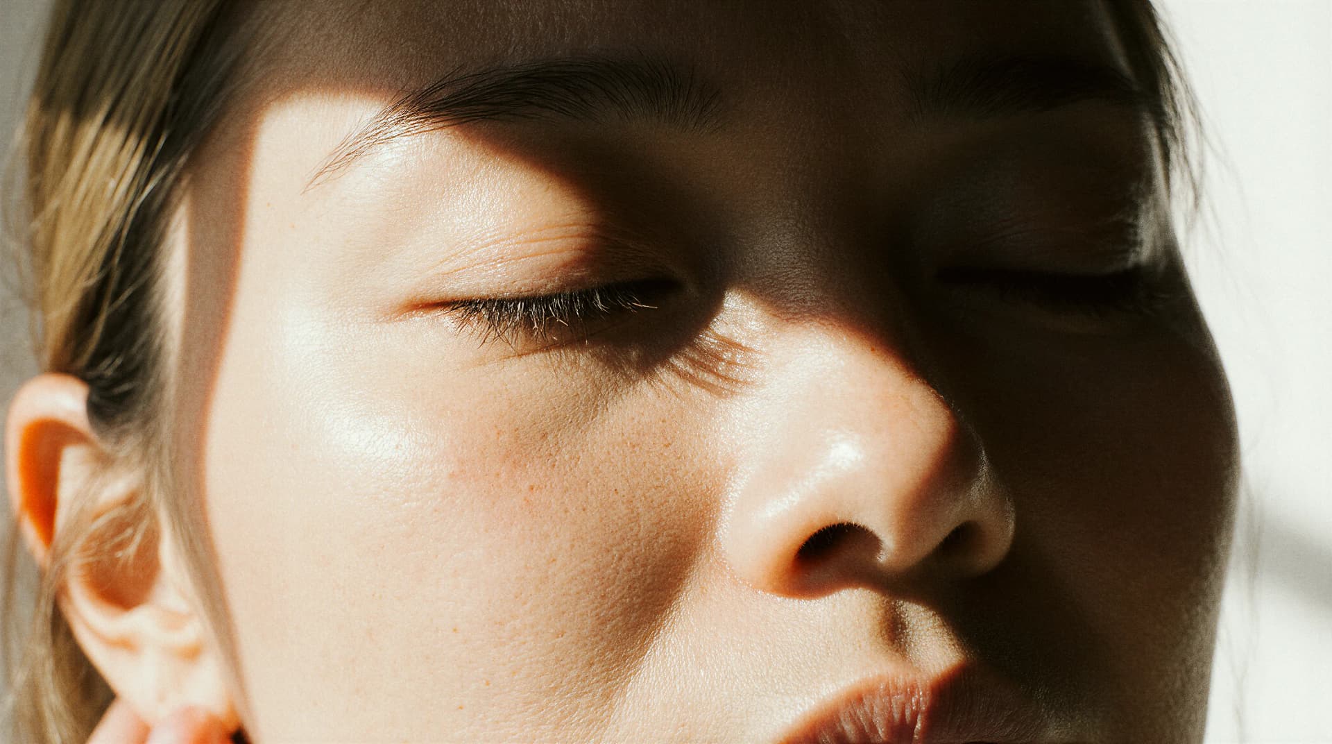 Close-up of a person's face with eyes closed, showing clear skin and soft shadows on the cheeks, nose, and forehead.